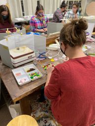 Masked crafters in a community art studio assembling colorful glass mosaic tiles at a workshop table with a kiln, trays, and tools.