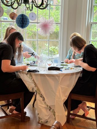 Cozy home craft party with four people at a sunlit dining table by a large window making jewelry and bead crafts with tools, wine glasses, and hanging paper pom-pom decorations