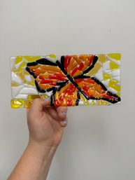 Handheld rectangular fused-glass mosaic panel depicting a vibrant orange, red and black butterfly on white and yellow background, held against a plain white wall — handmade glass art, stained-glass style.