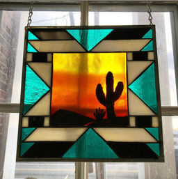 Southwestern-style stained-glass window panel showing an orange-yellow desert sunset with a silhouetted saguaro cactus in the center, framed by teal, white and black geometric glass, hanging by chains.