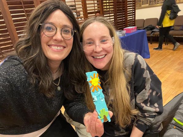 Two smiling craft workshop participants indoors holding a turquoise foam craft decorated with a yellow sunburst and green accents.