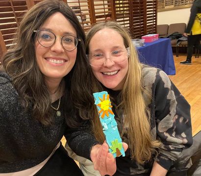 Two smiling craft workshop participants indoors holding a turquoise foam craft decorated with a yellow sunburst and green accents.