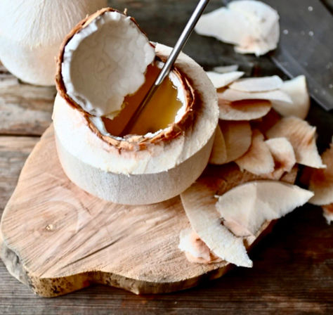 Peeled young coconut with the top cut off, metal straw and golden coconut water inside, coconut flesh shavings scattered on a wooden cutting board
