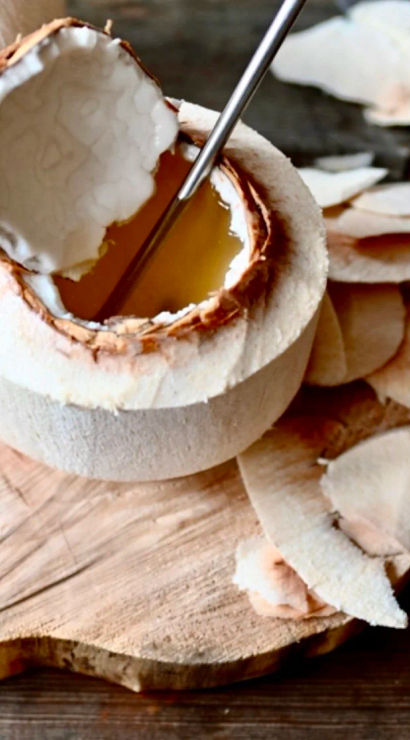 Peeled young coconut with the top cut off, metal straw and golden coconut water inside, coconut flesh shavings scattered on a wooden cutting board
