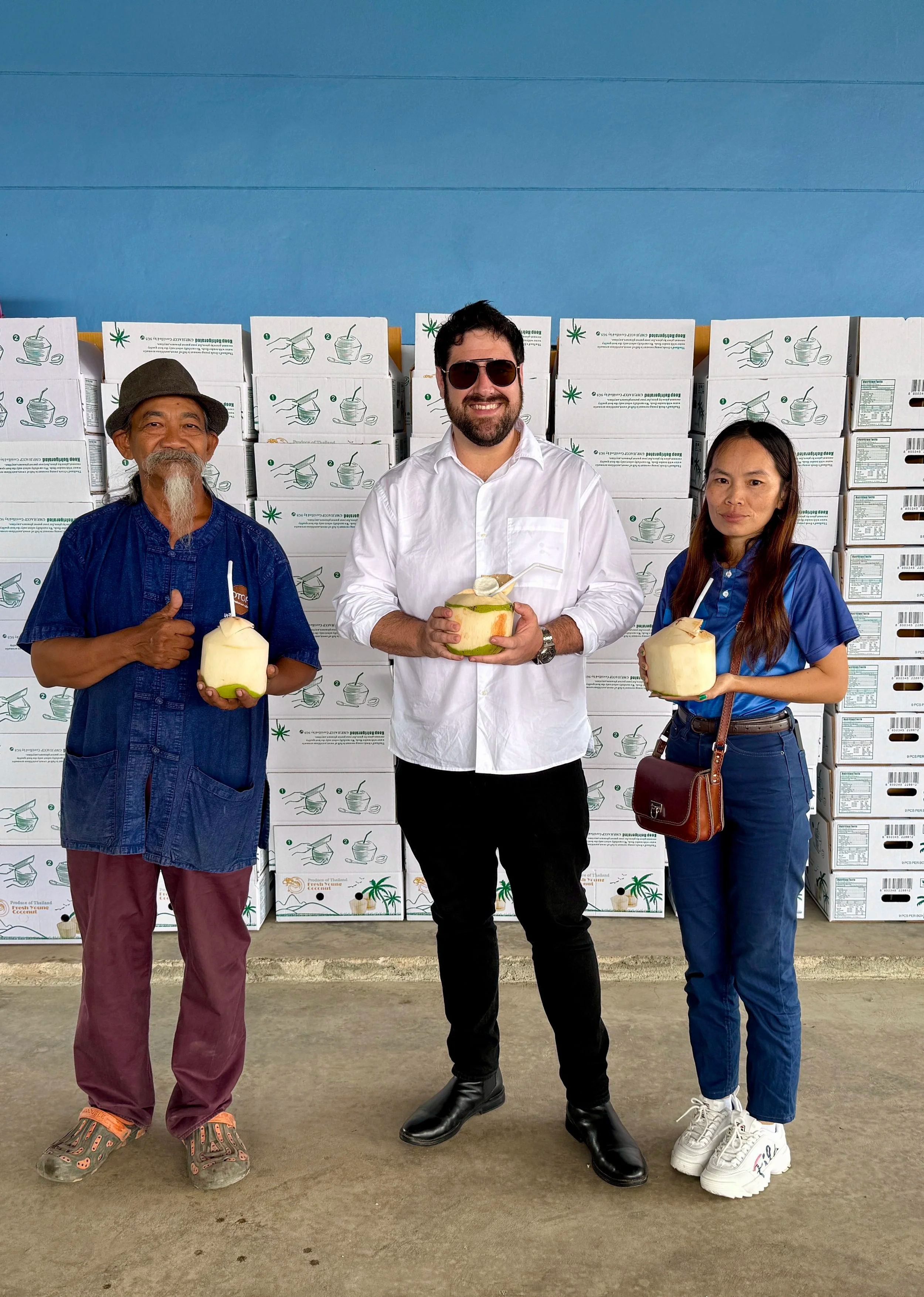 Three people pose in front of stacked produce boxes in a packing warehouse, each holding a fresh young coconut with a straw — casual, smiling group against a blue wall.