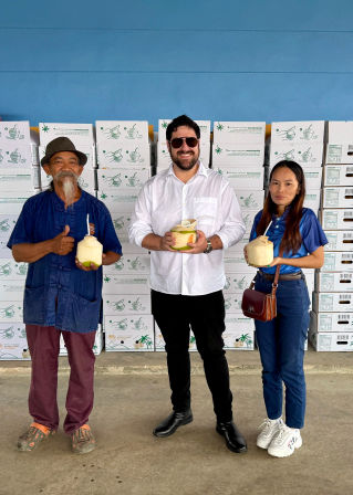 Three people pose in front of stacked produce boxes in a packing warehouse, each holding a fresh young coconut with a straw — casual, smiling group against a blue wall.