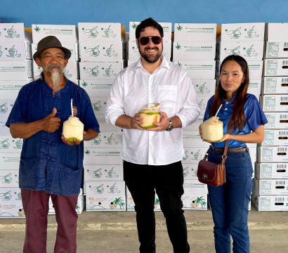 Three people pose in front of stacked produce boxes in a packing warehouse, each holding a fresh young coconut with a straw — casual, smiling group against a blue wall.