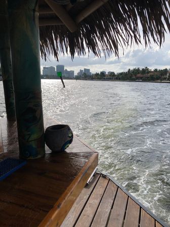 View from a tiki-hut deck over a sparkling waterway with a painted post and wooden dock, green channel markers, palm-lined shore and distant city skyline under a cloudy sky.