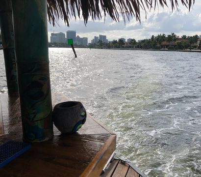 View from a tiki-hut deck over a sparkling waterway with a painted post and wooden dock, green channel markers, palm-lined shore and distant city skyline under a cloudy sky.