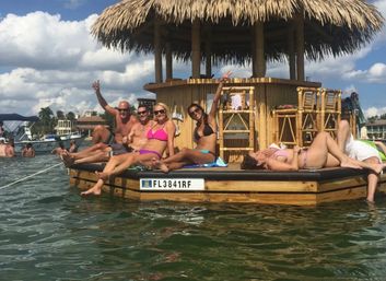 Group of friends in colorful swimsuits lounging and waving on a floating tiki bar with a thatched roof in sunny Florida coastal waters, boats and waterfront homes in the background.