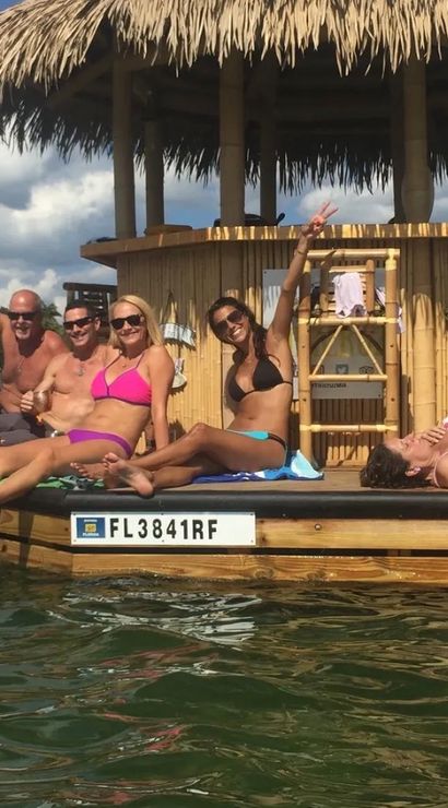 Group of friends in colorful swimsuits lounging and waving on a floating tiki bar with a thatched roof in sunny Florida coastal waters, boats and waterfront homes in the background.