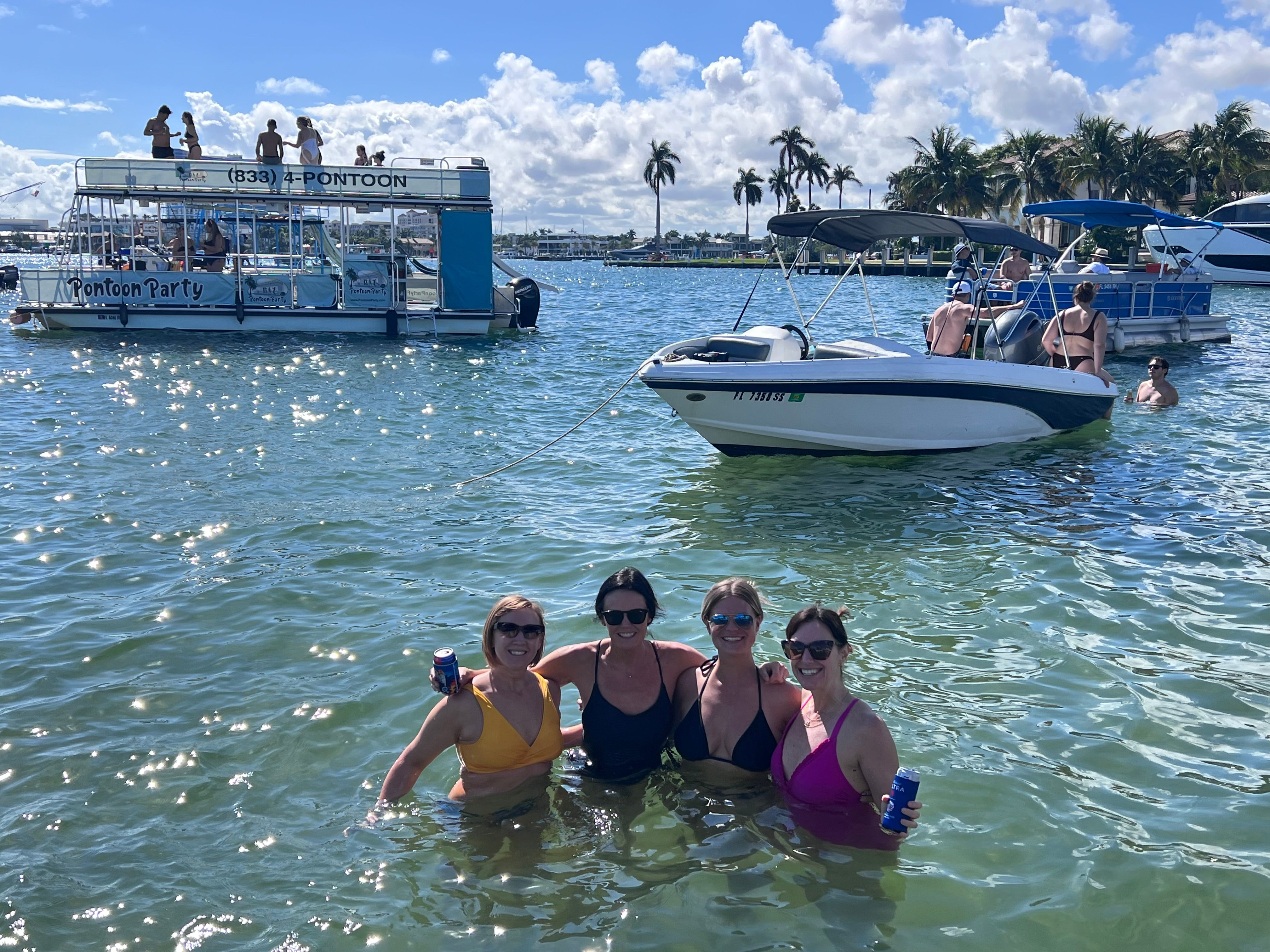 Four women in colorful swimsuits waist-deep in a sunny coastal bay, holding drinks and smiling near anchored boats and a party pontoon with palm trees and blue sky in the background.