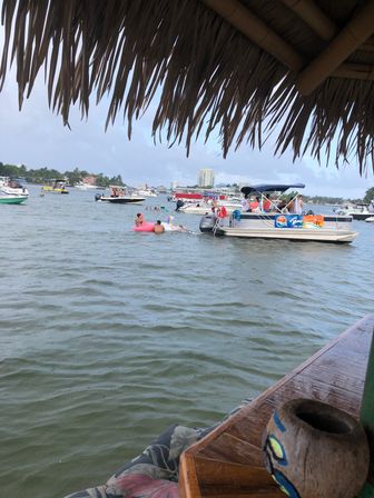 View from under a thatched palapa over a busy tropical bay with anchored boats and a pontoon party, people on a pink inflatable float, and skyline buildings on the distant shore.
