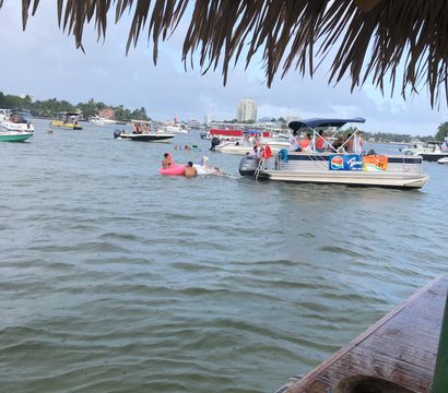 View from under a thatched palapa over a busy tropical bay with anchored boats and a pontoon party, people on a pink inflatable float, and skyline buildings on the distant shore.