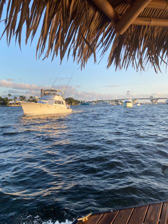 White sportfishing boat cruising past a marina at golden hour, viewed from a thatched-roof dock over rippling blue water with other boats and a bridge in the distance.