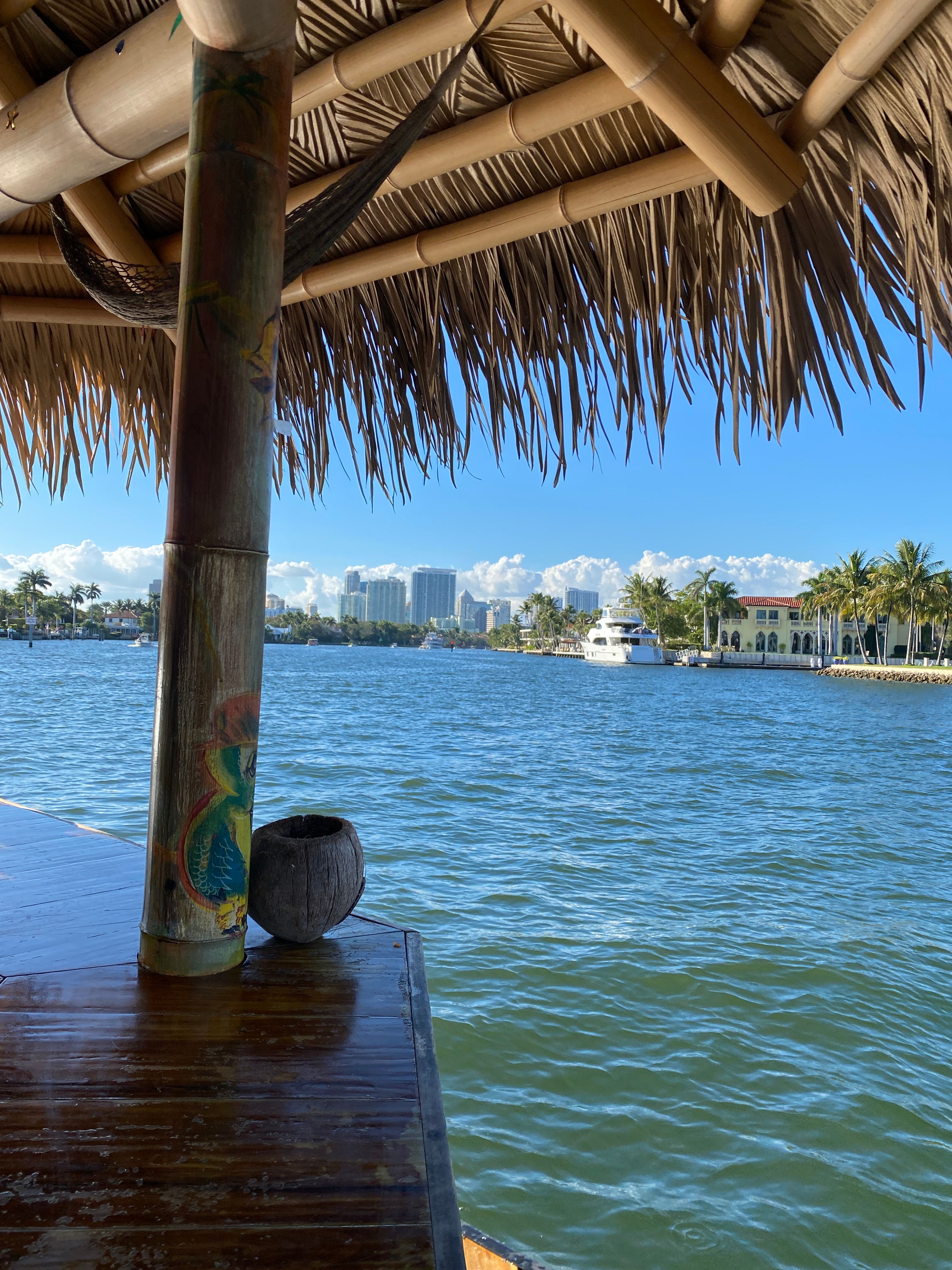 Foreground bamboo tiki hut post painted with a parrot and a coconut bowl, overlooking a turquoise bay with a palm-lined waterfront, moored yacht and city skyline under a bright blue sky.
