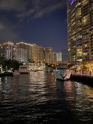 Waterfront marina at night with glowing high-rise condos, docked yachts, a lively boardwalk, and shimmering reflections on the water.