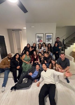 Large group of friends smiling and posing for a lively living-room group photo in a modern home, seated on couches and the floor near a staircase and framed wall art.