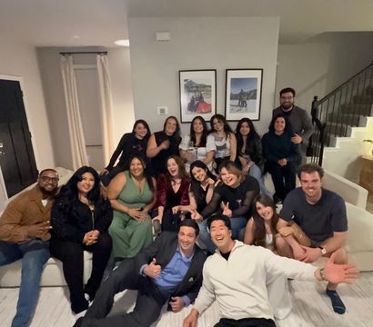 Large group of friends smiling and posing for a lively living-room group photo in a modern home, seated on couches and the floor near a staircase and framed wall art.
