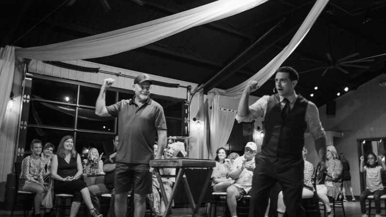 Black-and-white shot of a live indoor performance: a headset-wearing entertainer and a casually dressed participant raise fists on stage while a smiling audience applauds beneath draped fabric in a cozy event space.