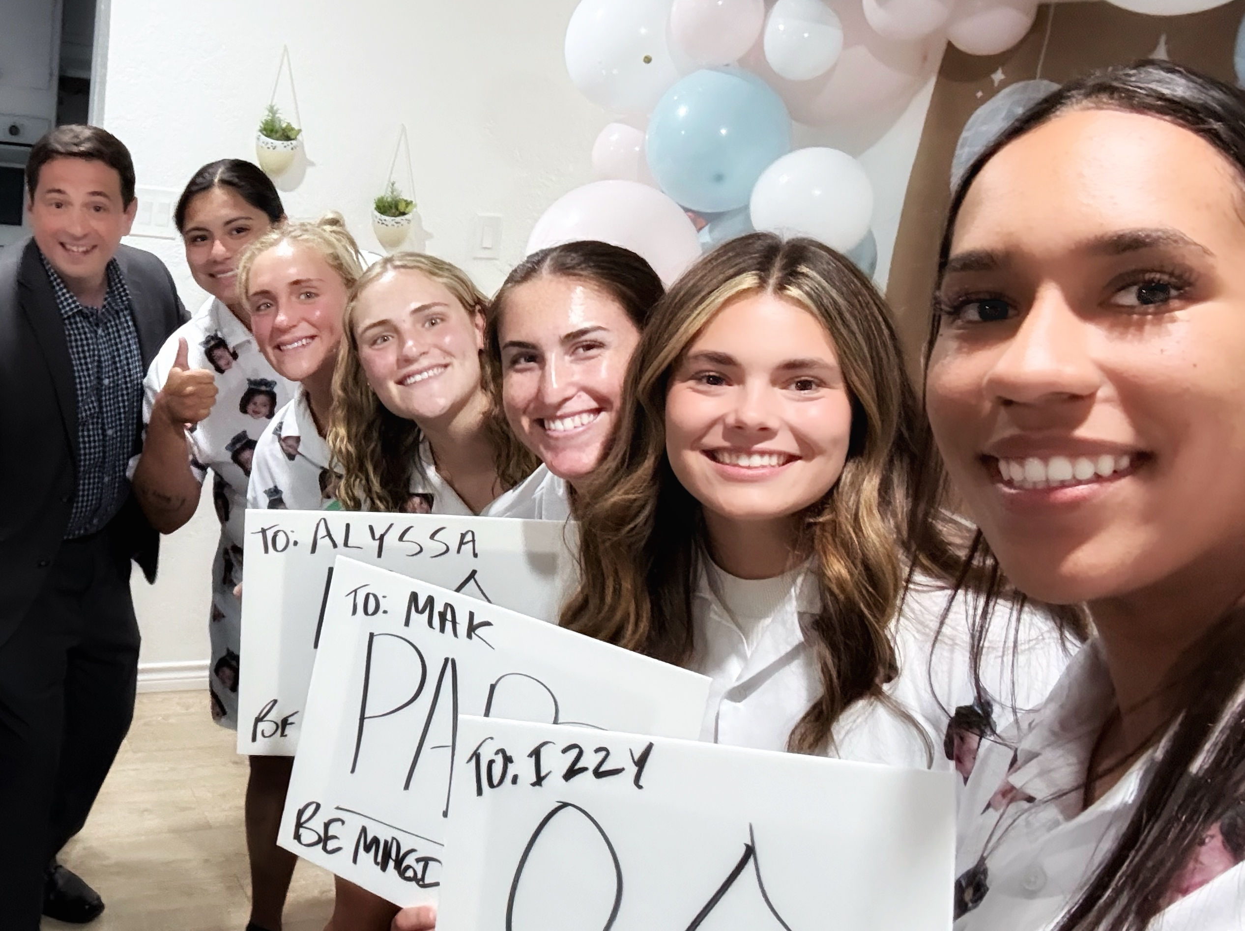 Group selfie of seven smiling friends at an indoor celebration holding handmade signs with a pastel balloon garland and a thumbs-up, casual white shirts and festive atmosphere.