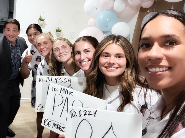 Group selfie of seven smiling friends at an indoor celebration holding handmade signs with a pastel balloon garland and a thumbs-up, casual white shirts and festive atmosphere.