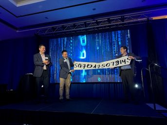 Three suited presenters laugh while holding an upside-down banner on a conference stage with a glowing blue LED screen showing vertical binary code.