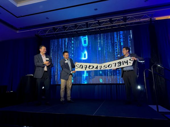 Three suited presenters laugh while holding an upside-down banner on a conference stage with a glowing blue LED screen showing vertical binary code.