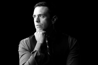 Black-and-white studio portrait of a pensive man in a dress shirt, vest and tie, chin resting on hand, dramatic side lighting against a black background.