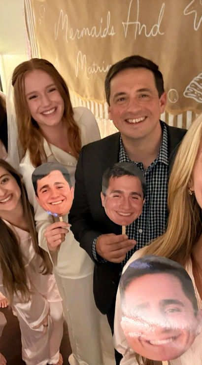 Smiling group selfie at a pajama party — friends in matching satin pajamas and one person in a jacket holding face-cutout props in front of a decorated party backdrop