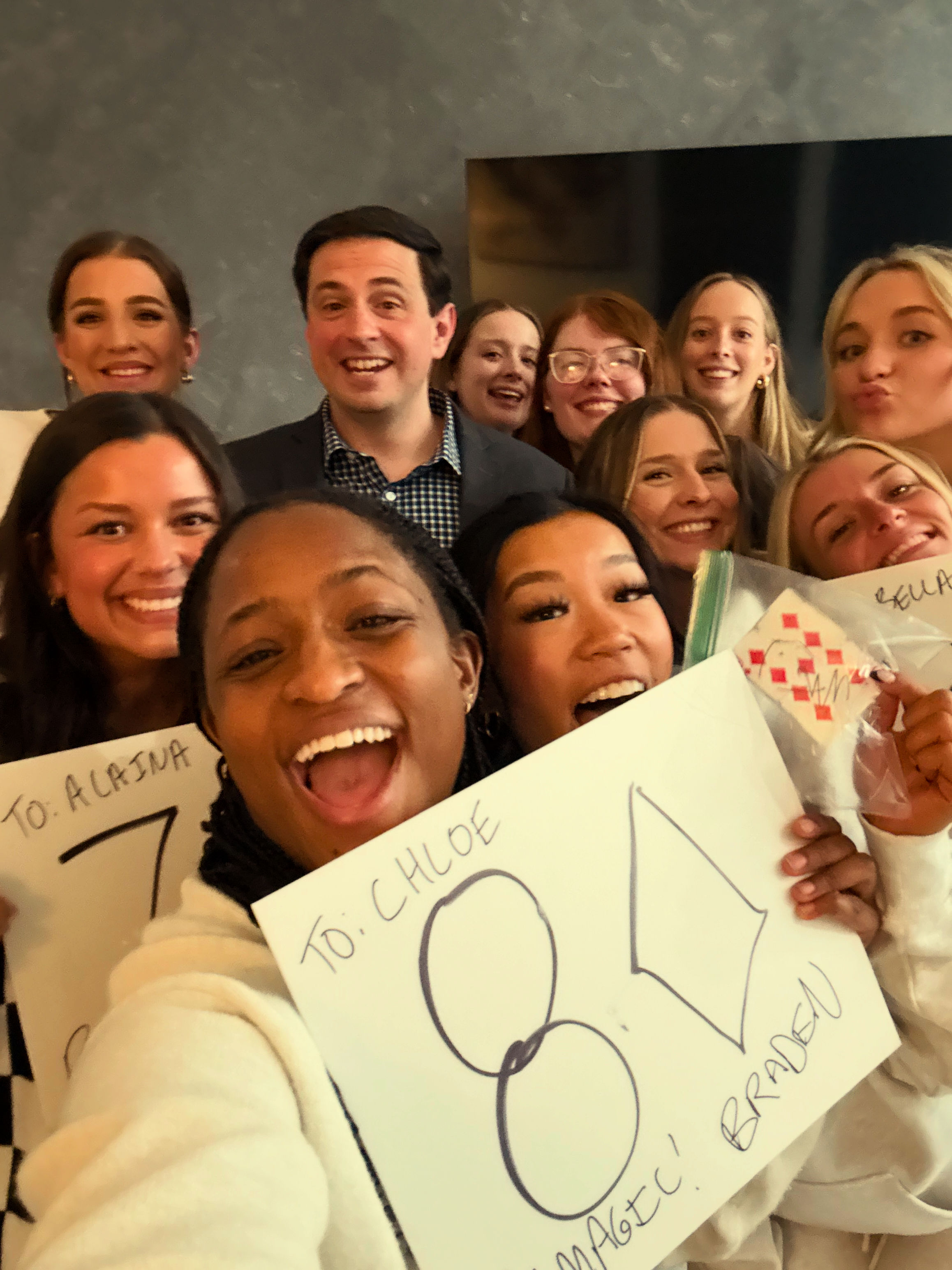 Cheerful indoor group selfie of smiling young adults crowded together, many holding handmade signs featuring large "80" and celebratory messages.