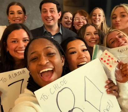 Cheerful indoor group selfie of smiling young adults crowded together, many holding handmade signs featuring large "80" and celebratory messages.