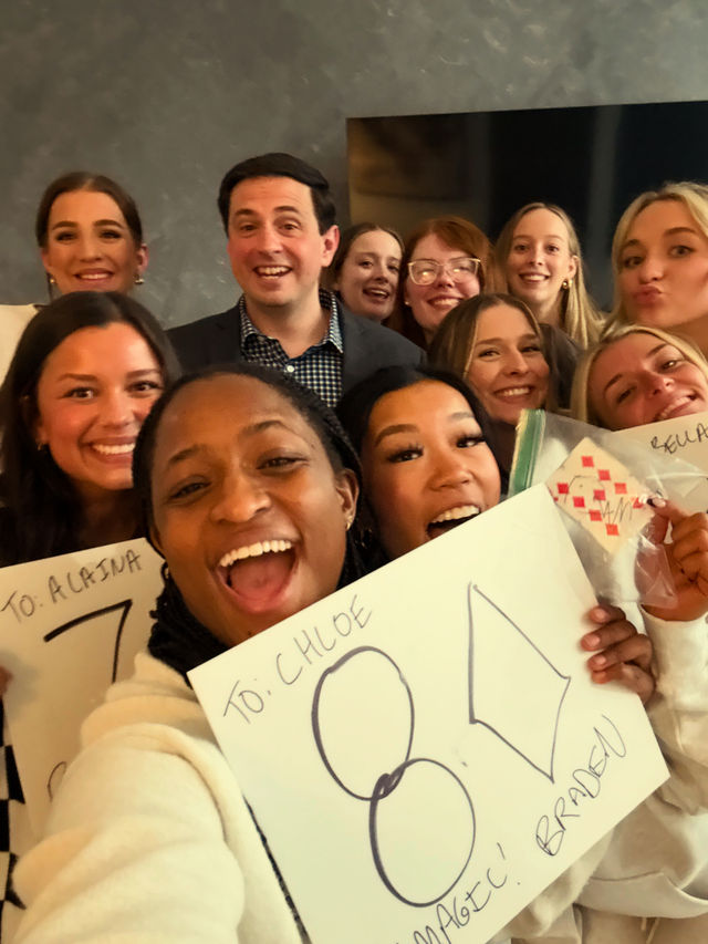 Cheerful indoor group selfie of smiling young adults crowded together, many holding handmade signs featuring large "80" and celebratory messages.