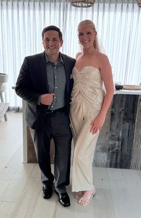 Smiling couple in formal attire posing in a light-filled modern venue with sheer curtains — man in a dark suit and checked shirt, woman in a strapless cream bridal gown with veil and strappy heels.