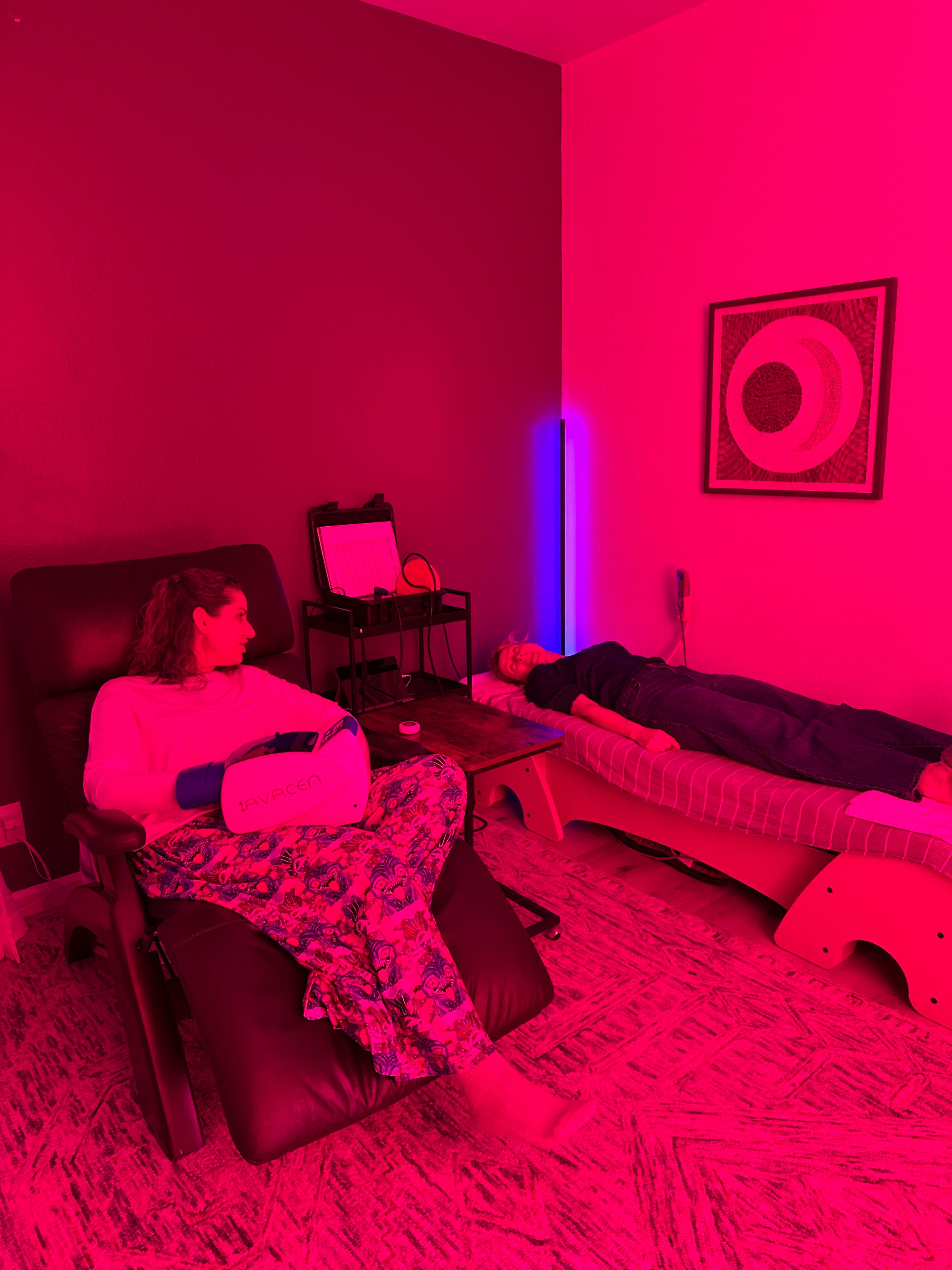 Two people in a pink-lit wellness room — one reclining in a chair using a handheld therapy device, the other resting on a cushioned treatment table, with a neon blue floor lamp and circular modern wall art.