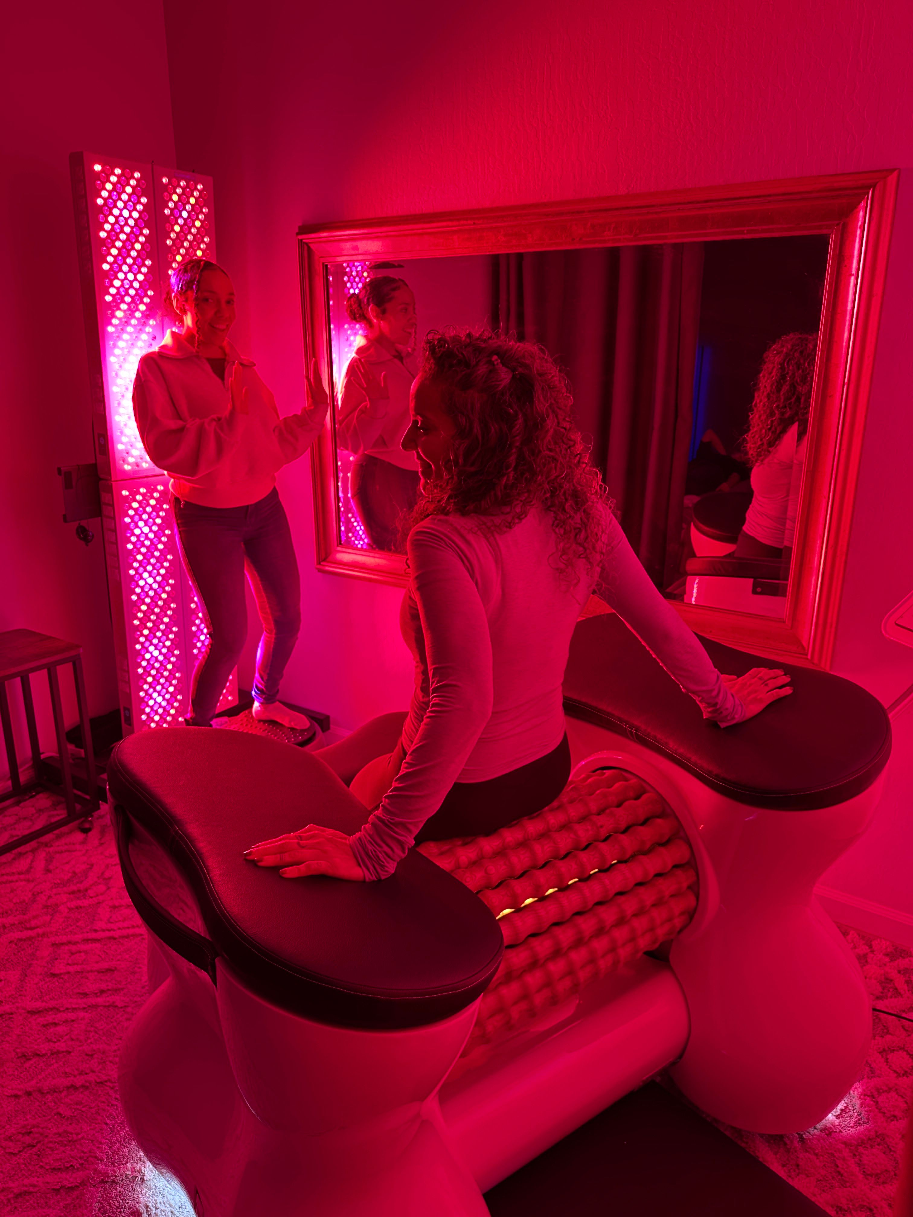 Woman using a red-light therapy roller machine in a pink-lit spa treatment room with LED panels and mirror reflection