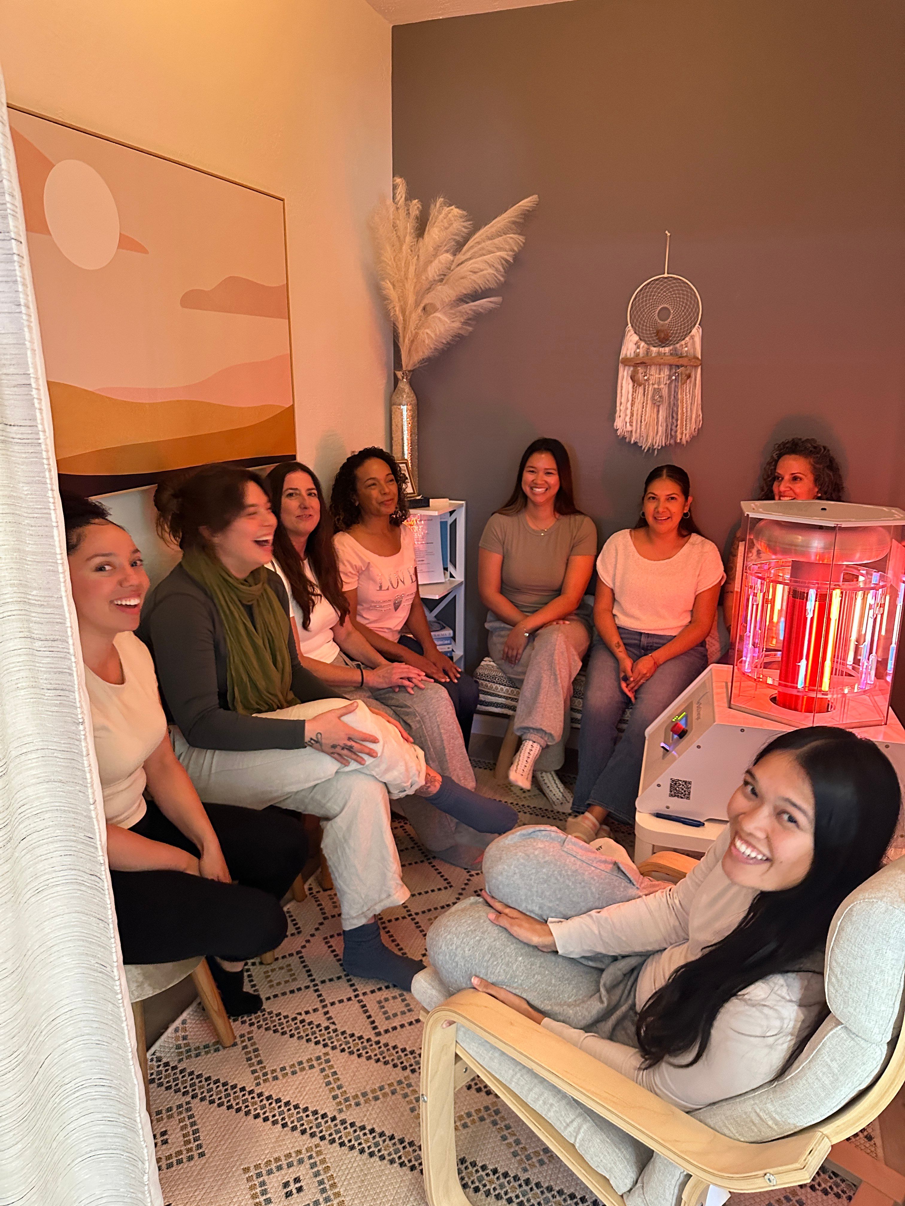 Group of smiling women seated in a cozy neutral-toned wellness studio around a red light therapy device, with boho decor like pampas grass and a dreamcatcher.