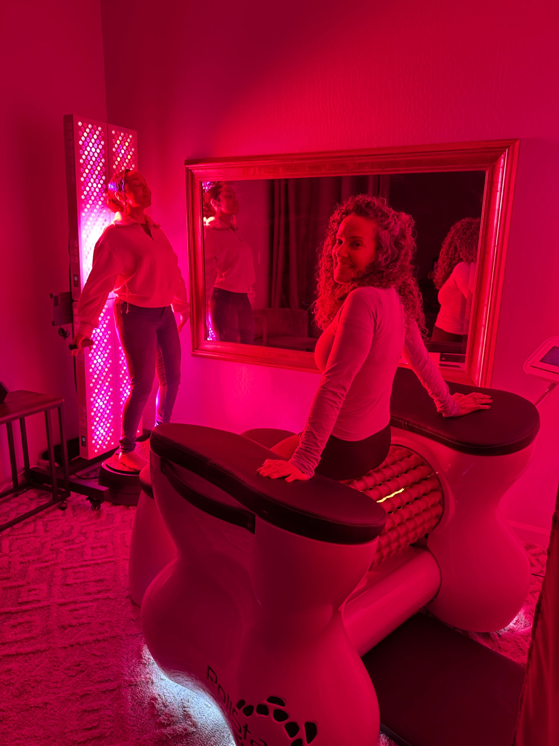 Two people in a glowing pink-red LED light therapy spa room — one seated on a rolling massage/lymphatic machine and another standing on a platform by a vertical infrared LED panel with their reflections in a wall mirror.