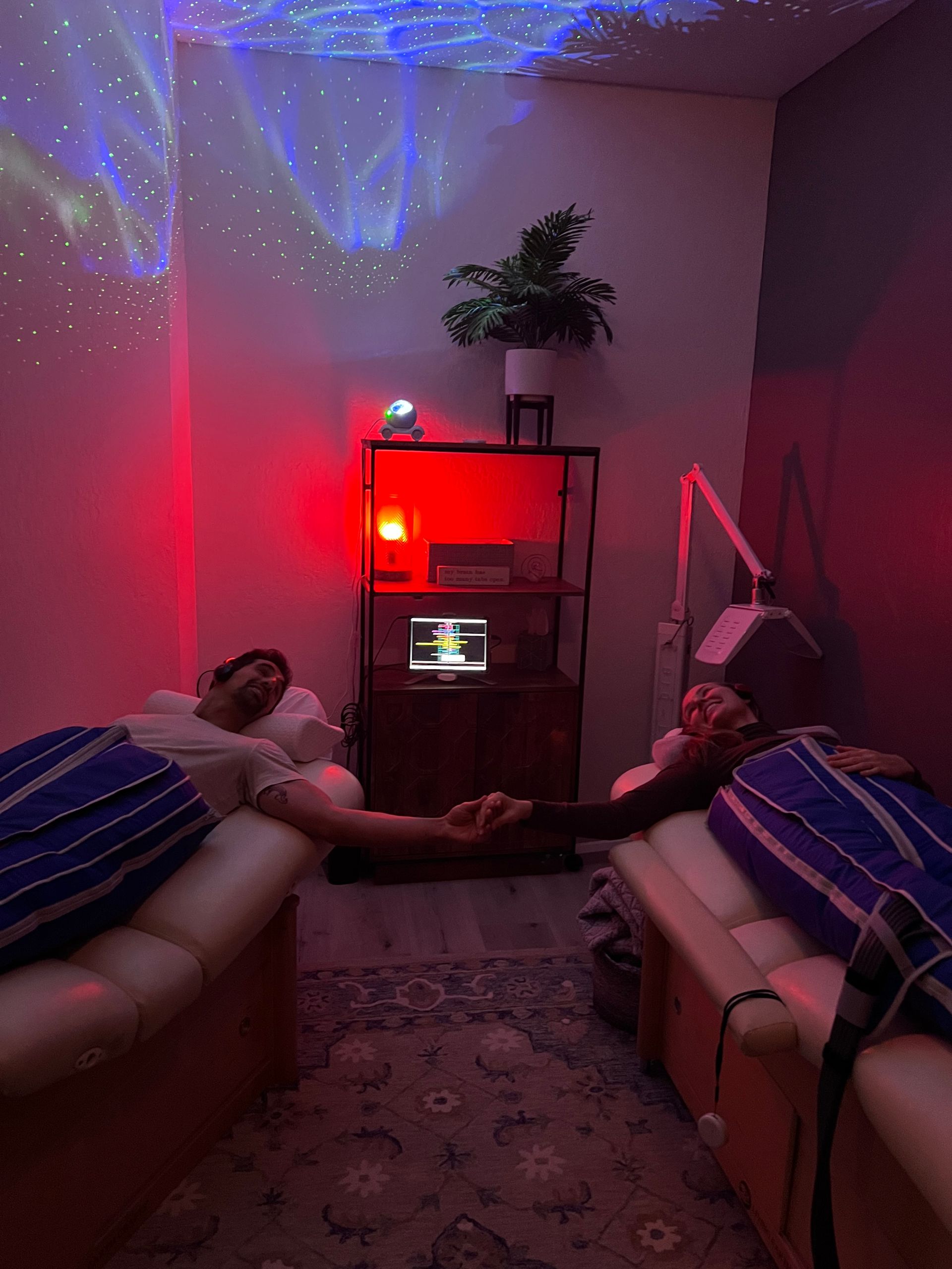 Two people holding hands during paired leg-compression therapy on lounge chairs in a cozy relaxation room with red mood lighting and a starry ceiling projector.