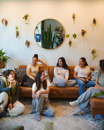 Cozy indoor gathering of friends laughing and chatting, six women relaxing on a tan leather sofa and patterned rug surrounded by hanging glass plant vases and a round wall mirror reflecting tall greenery
