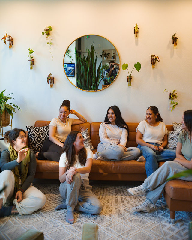 Six women in casual loungewear chatting and laughing in a cozy living room with a brown leather sofa, indoor plants, and a round wall mirror.