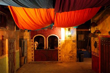 Cozy colorful courtyard with oversized orange fabric canopy overhead, arched service window framed by decorative tiles, warm lantern glow on weathered plaster walls, rustic doors and woven baskets.