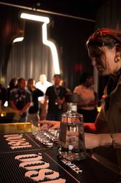 Bartender preparing tequila shots on a dim cocktail bar counter with a clear liquor bottle, lined-up shot glasses and a neon bottle outline above a crowd — lively nightlife scene.