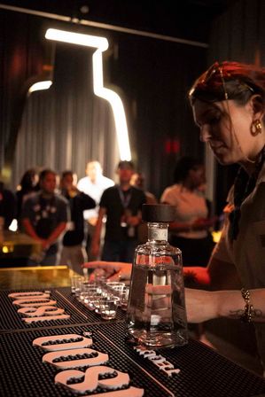 Bartender preparing tequila shots on a dim cocktail bar counter with a clear liquor bottle, lined-up shot glasses and a neon bottle outline above a crowd — lively nightlife scene.