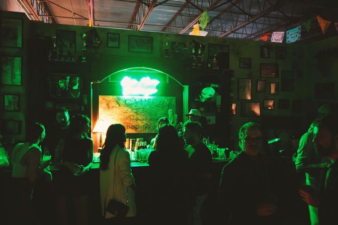 Neon-lit nightlife scene: dim indoor bar with a bright green neon sign above a wooden counter, silhouetted patrons chatting and sipping drinks under colorful hanging banners.