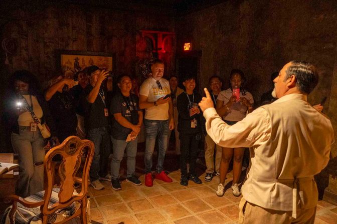Tour guide addressing a small group of visitors in a dimly lit historic room; guests record on phones and cameras, an ornate wooden chair in the foreground and a glowing exit sign in the background.