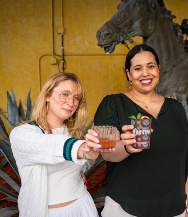 Two smiling women toasting cocktails on an outdoor patio with agave plants and a large horse sculpture in the background.