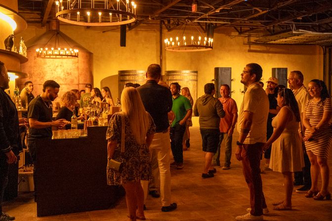 Crowded rustic wine tasting room with warm amber lighting, circular candle chandeliers, wooden barrels and a busy bar where patrons line up for drinks.