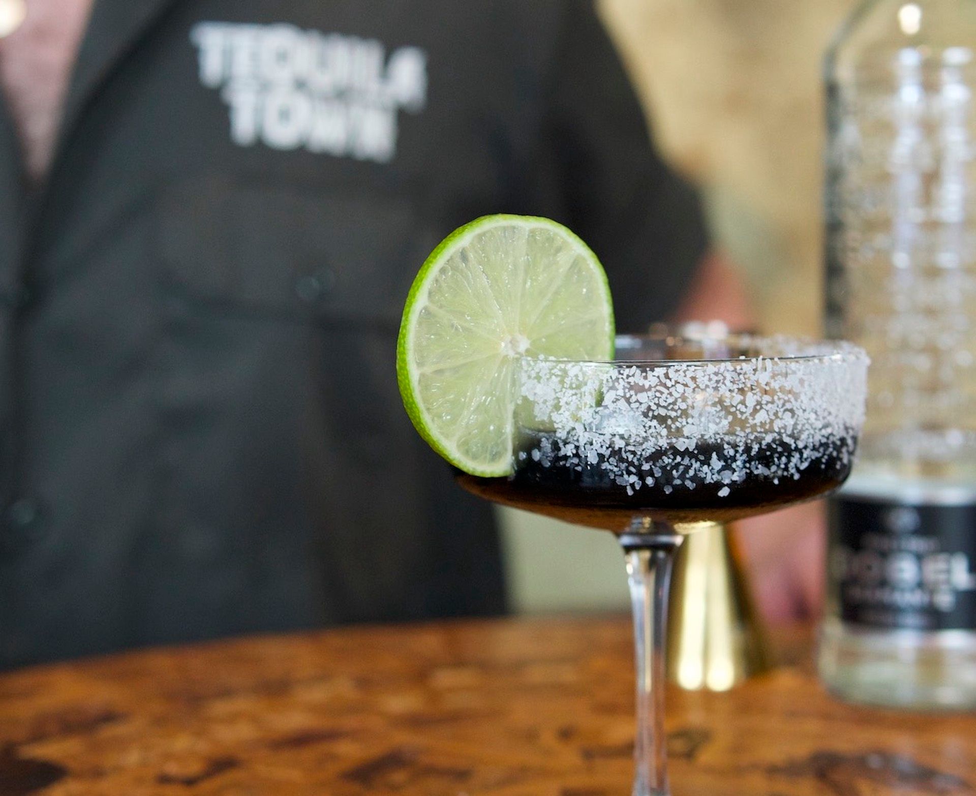 Dark, salt‑rimmed cocktail in a coupe glass with a zesty lime wheel garnish on a wooden bar, blurred bartender and bottle in the background.