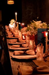 Cozy candlelit long wooden table set for dinner with folded linen napkins, textured glass tumblers, wine bottles and white floral centerpieces under warm ambient lighting
